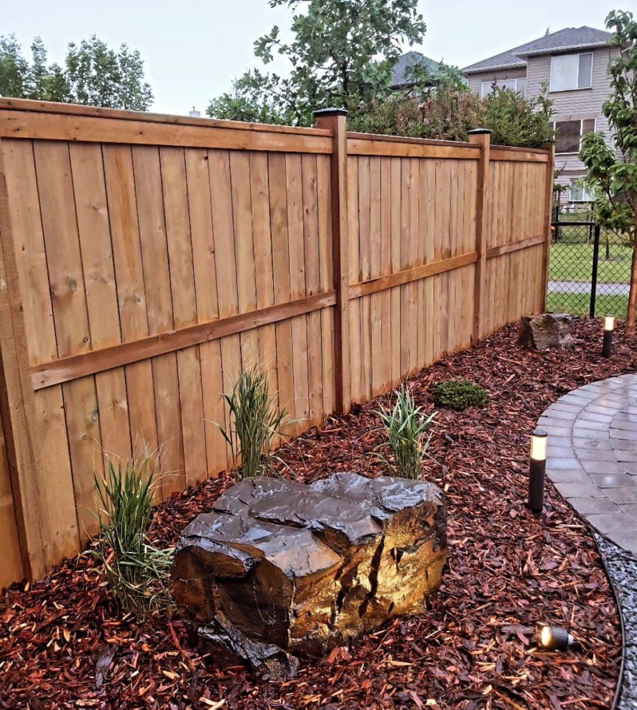 Dark reddish-brown bark mulch covers a garden bed along a wooden fence with plants and rocks.