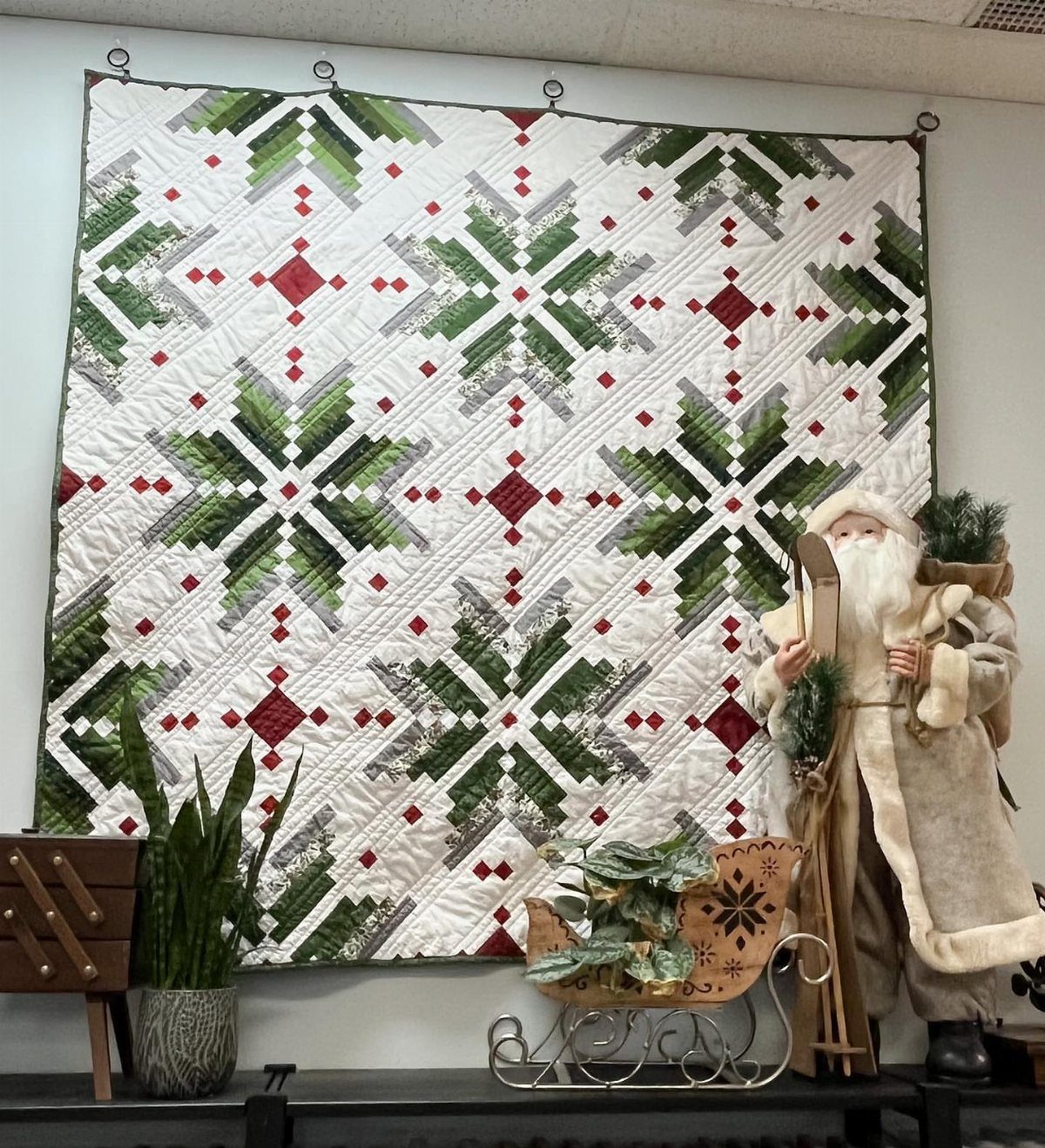 A white quilt with green and red star patterns hanging on a wall above festive decorations.