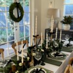 A festive dining table set with green napkins, gold cutlery, and a greenery centerpiece with tall candles.