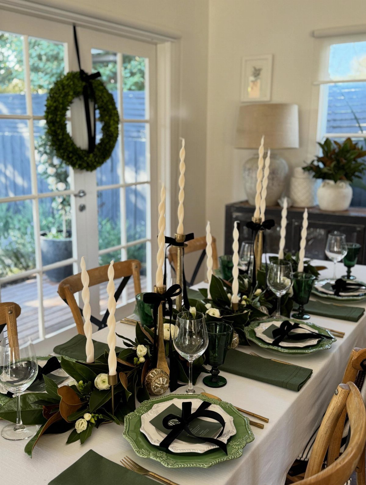 A festive dining table set with green napkins, gold cutlery, and a greenery centerpiece with tall candles.