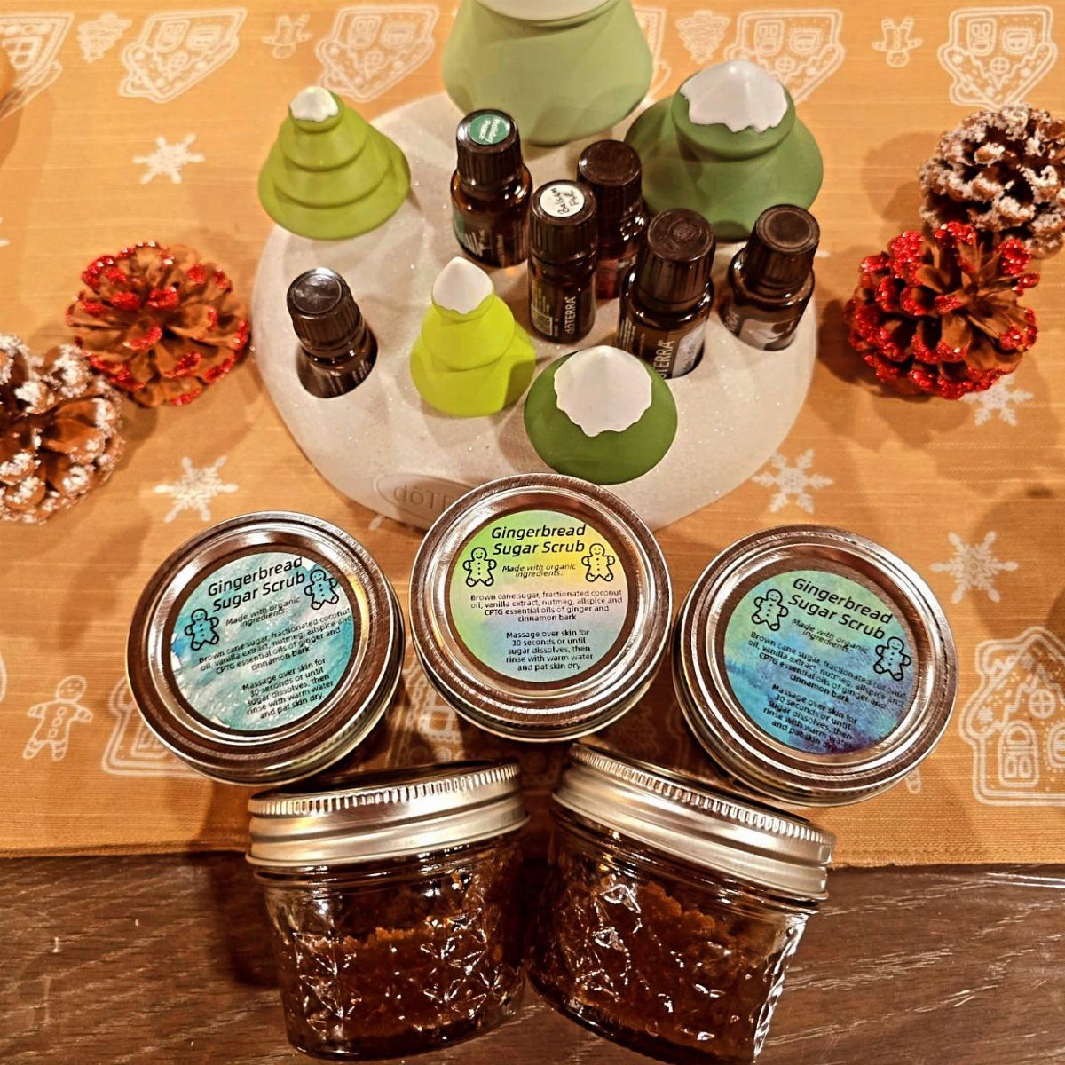 Four jars of gingerbread sugar scrub arranged with essential oils and decorative ceramic trees.