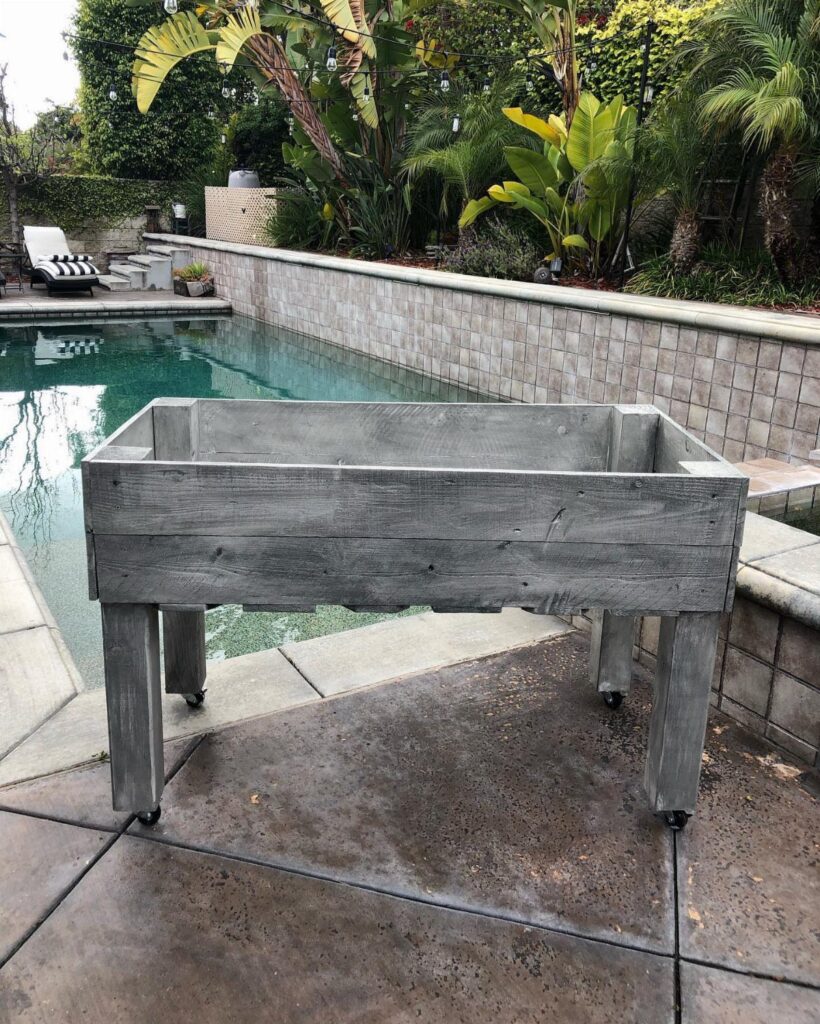 Grey wooden planter box with wheels sitting on a patio next to a swimming pool.