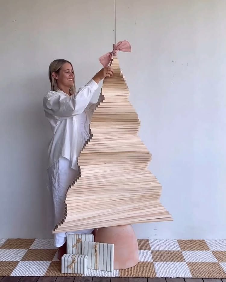 A woman stands beside a large decorative Christmas tree constructed from stacked wooden slats.